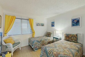 Ein Zimmer mit zwei Einzelbetten, einem Sessel, einem Tisch und einem Vorhangfenster.