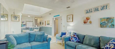 A living room with two blue sofas, a ceiling fan, and a wall sign that reads 'SEAS THE DAY'.