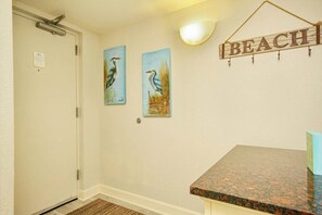 Un couloir avec une porte, deux tableaux représentant des oiseaux accrochés au mur, une enseigne murale sur laquelle on peut lire « PLAGE », et un comptoir en granit.