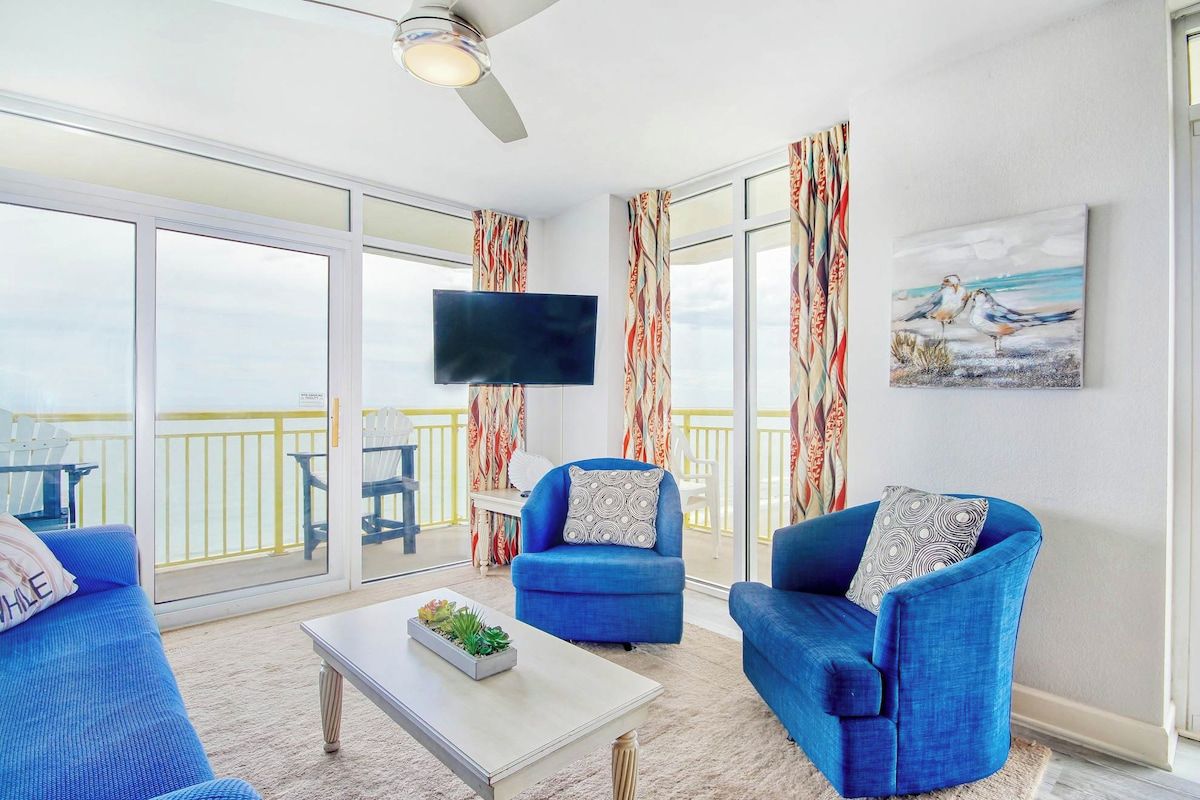 A room with a large sliding glass door leading to a balcony, a flat-screen TV mounted on the wall, and two blue upholstered chairs with patterned cushions.