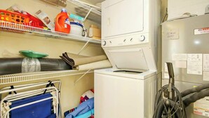 A laundry area with a stacked washer and dryer, shelves with cleaning supplies, and a cart with folded clothes. - Bay Watch Resort 1740 (North Myrtle Beach)