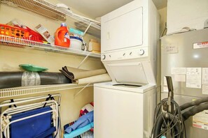 A laundry area with a stacked washer and dryer, shelves with cleaning supplies, and a cart with folded clothes. - Bay Watch Resort 1740 (North Myrtle Beach)