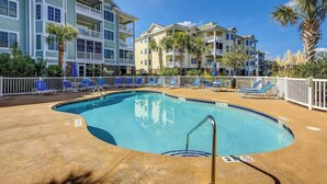 A swimming pool with lounge chairs and a poolside fence, surrounded by multi-story buildings with balconies. - Luster Leaf Unit 201 (Myrtle Beach)