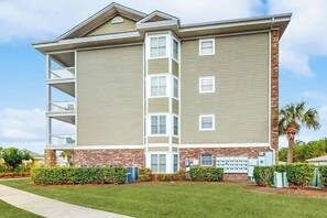 A multi-story building with a brick base and beige siding, featuring a central staircase, balconies, and a landscaped front yard. - Luster Leaf Unit 201 (Myrtle Beach)