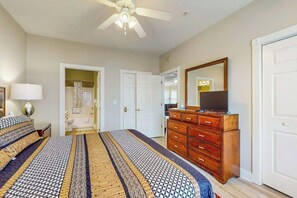 Une chambre à coucher avec un lit, une commode, un ventilateur de plafond et une salle de bain visible à travers une porte ouverte.