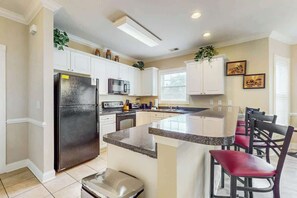 A modern kitchen with white cabinets, a stainless steel refrigerator, a microwave, and a granite countertop island with bar stools. - Luster Leaf Unit 201 (Myrtle Beach)