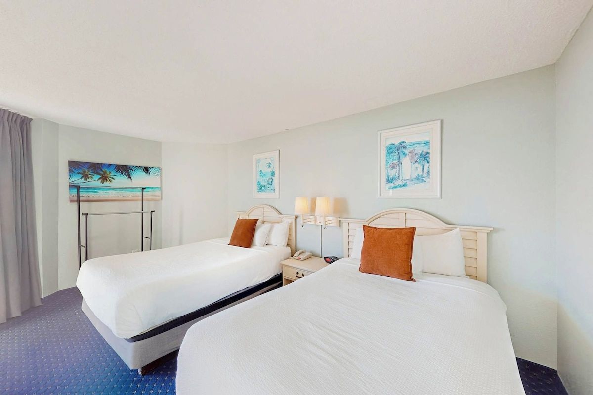 A hotel room with two beds, a nightstand, a lamp, and artwork on the wall.