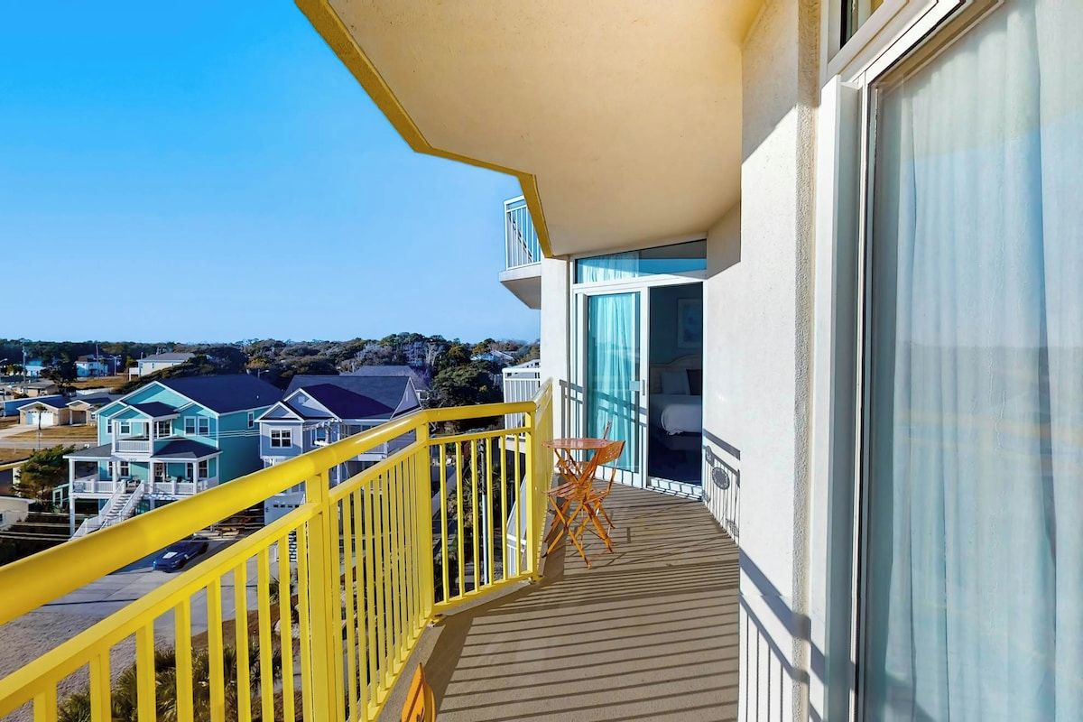 Een balkon met een geel hekwerk, een klein tafeltje en een uitzicht op huizen in de verte.