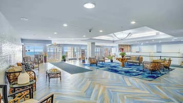Eine geräumige Lobby mit einem Fischgrätenmuster-Boden, Sitzbereichen mit Korbmöbeln und Blick auf das Meer durch große Fenster.