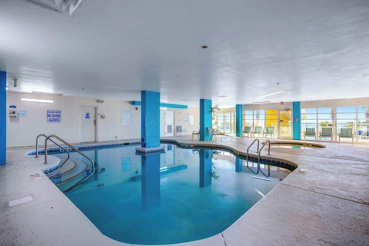Une piscine intérieure à l’eau claire, avec des murs carrelés de bleu et de blanc, et un espace baigné de lumière où l’on peut s’asseoir.