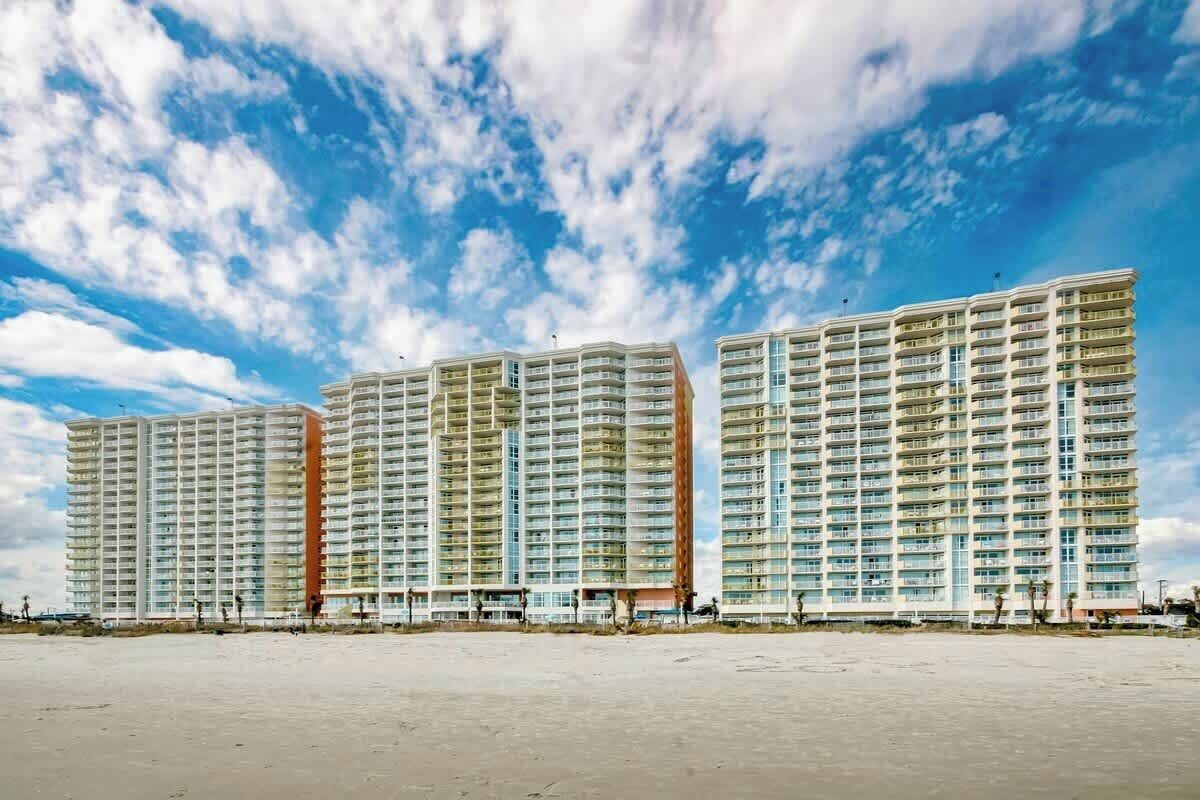 Three high-rise residential buildings with multiple balconies, situated on a flat terrain.