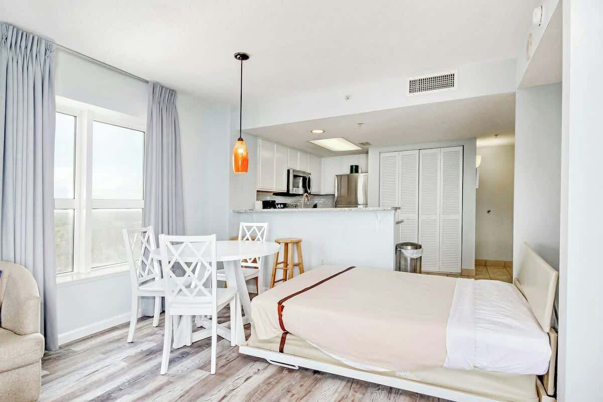 A compact hotel room with a bed, a dining table with chairs, a kitchenette, and a window with curtains.