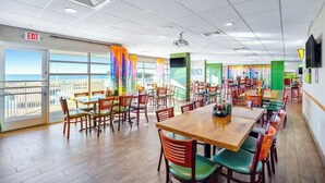 A spacious indoor dining area with multiple tables and chairs, large windows with curtains, and an exit sign. - Bay Watch Resort 1741 (North Myrtle Beach)