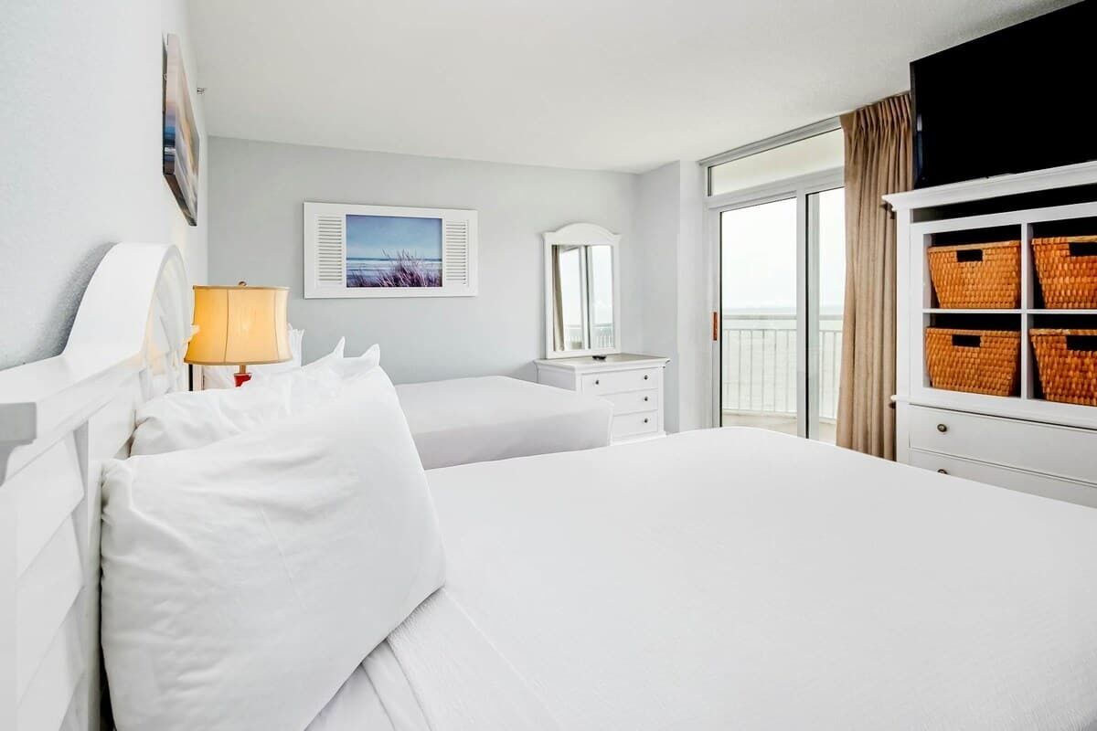 Habitación de hotel con una cama grande, una mesita de noche con lámpara, cómoda, espejo y ventana con cortinas.