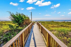 Ein Holzsteg führt zu einem Strand, mit blauem Himmel und weiter Meerblick.