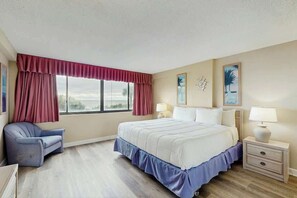 A hotel room with a bed, a chair, a nightstand, and a window with curtains. - Carolina Dunes Unit 102 (Myrtle Beach)