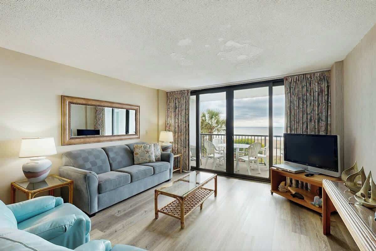 A hotel room with a sofa, a coffee table, a TV on a wooden stand, and a balcony with a view.