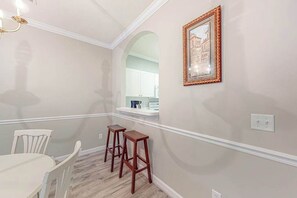 Ein Essbereich mit einem weißen Tisch und Stühlen, zwei hölzernen Hocker, einem gerahmten Bild an der Wand und einer bogenförmigen Durchgangstür zu einer Küche.