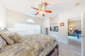 Ein Schlafzimmer mit einem Bett, einem Deckenventilator, einem Fernseher und einem Flur, der in einen weiteren Raum führt.
