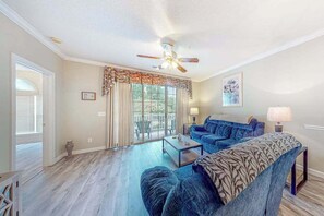 Ein Wohnzimmer mit Deckenventilator, einer Couch, einem Couchtisch und Blick durch eine Glastür auf einen Außenbereich.