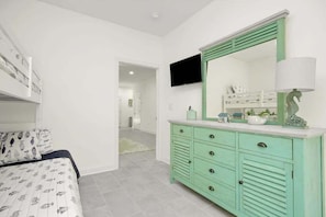 A bedroom with a bed, a green dresser, a television, and a hallway leading to another room. - Grace by the Sea Retreat (Atlantic Beach)
