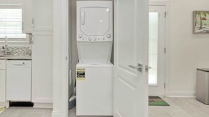 A compact kitchen with a dishwasher, a white refrigerator, and a door leading to another room. - Grace by the Sea Retreat (Atlantic Beach)