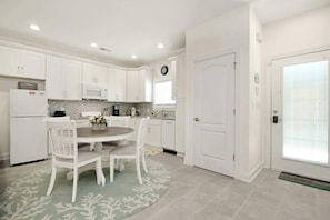 A kitchen with white cabinets, a microwave, a clock, and a dining area with a table and chairs. - Grace by the Sea Retreat (Atlantic Beach)