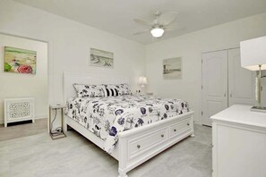 A bedroom with a bed, a nightstand, a lamp, a ceiling fan, and artwork on the walls. - Grace by the Sea Retreat (Atlantic Beach)