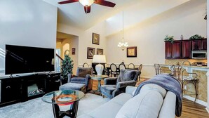A living room with a television, sofa, chairs, a glass coffee table, and a ceiling fan. - Clearwater Bay Unit 1012 (North Myrtle Beach)