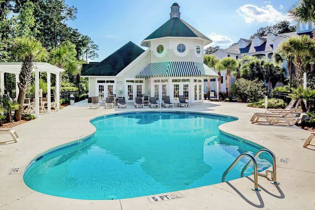 A swimming pool with lounge chairs and a gazebo, surrounded by palm trees and a building with a green roof.
