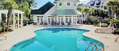 A swimming pool with lounge chairs and a gazebo, surrounded by palm trees and a building with a green roof.