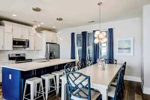 A modern kitchen with white cabinets, a central island with bar stools, and a dining area with blue chairs. - Blue Wave Beach Retreat (Murrells Inlet)