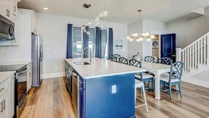 A modern kitchen with a central island, white cabinetry, and a dining area with blue chairs. - Blue Wave Beach Retreat (Murrells Inlet)