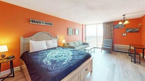 A hotel room with a bed, a desk, a chair, and a sofa. There is a sign on the wall that reads 'BEACH HOUSE'. - Sands Ocean Club 1101 Fruit Cakes (Myrtle Beach)