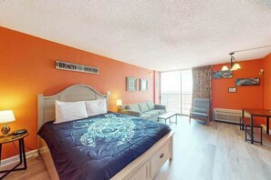 A hotel room with a bed, a desk, a chair, and a sofa. There is a sign on the wall that reads 'BEACH HOUSE'. - Sands Ocean Club 1101 Fruit Cakes (Myrtle Beach)