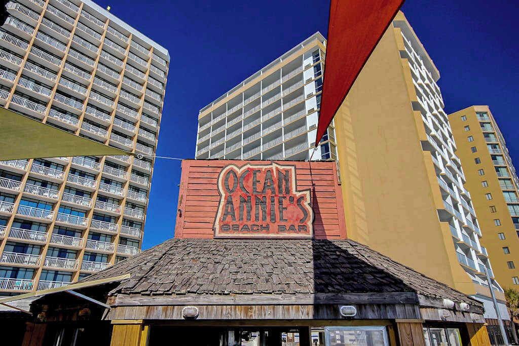 Um edifício com uma placa indicando 'Ocean Annie's Beach Bar' e vista de edifícios altos ao fundo.