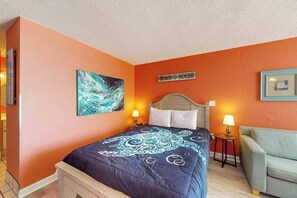 A bedroom with a bed, a nightstand, a chair, and a wall sign. - Sands Ocean Club 1101 Fruit Cakes (Myrtle Beach)