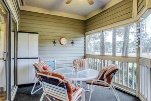 A covered outdoor seating area with a round table and chairs, a clock on the wall, and a view of trees through the window. - Tidewater 4214 Condo (North Myrtle Beach)