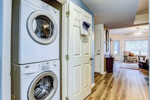 A laundry room with stacked washers and dryers, a door with a towel rack, and a living area with a sofa and a lamp. - Tidewater 4214 Condo (North Myrtle Beach)