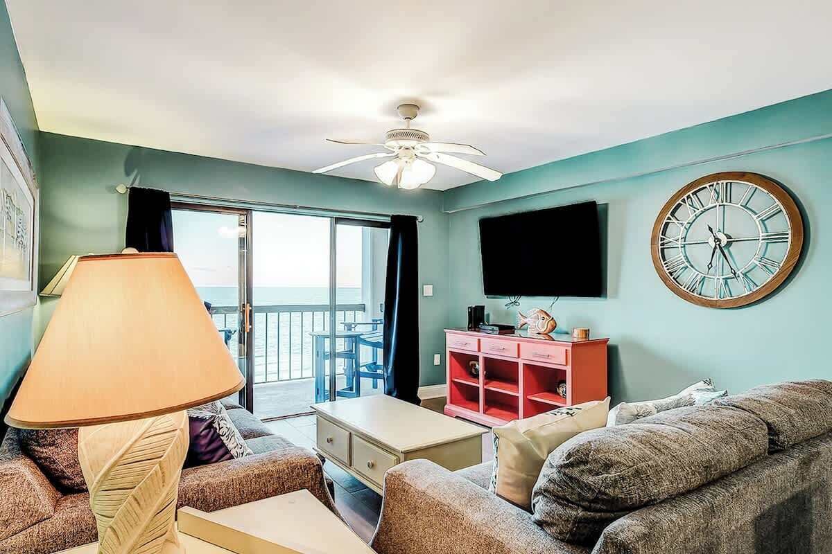 A living room with a ceiling fan, a television, a clock, and a view of the outdoors.