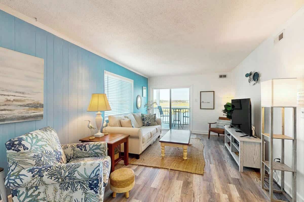 Un salon comprenant un canapé, une table basse et un meuble pour la télévision. Il y a une fenêtre avec des stores, une lampe et un tableau accroché au mur.