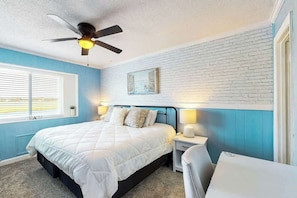 Une chambre à coucher comprenant un lit, des tables de chevet, un ventilateur de plafond, une fenêtre avec des stores et un tableau accroché au mur.