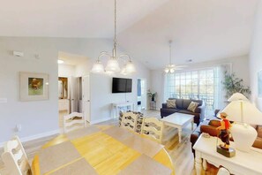 A modern living room with a dining area, a sofa set, a TV mounted on the wall, and a large window providing natural light. - Ironwood 1532 2 BR Condo (North Myrtle Beach)