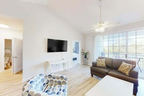 A modern living room with a flat-screen TV, a sofa, a coffee table, and a large window offering a view. - Ironwood 1532 2 BR Condo (North Myrtle Beach)