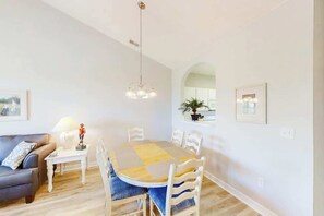 A dining area with a round table, chairs, a hanging light fixture, a sofa, and a window with a view. - Ironwood 1532 2 BR Condo (North Myrtle Beach)