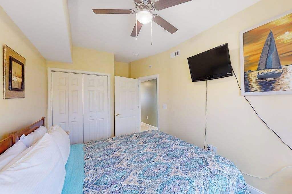 Un dormitorio con cama, ventilador de techo, televisión y un cuadro en la pared.