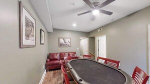 Una habitación con mesa de billar, sillas rojas, ventilador de techo y cuadros en la pared.