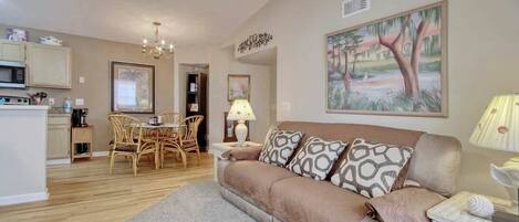 A living room with a sofa, a dining table with chairs, a kitchen area, and a painting on the wall.
