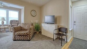 A living room with a sofa, a television on a cabinet, a chair, a clock on the wall, and a door. - Home Away From Home Unit J (Surfside Beach)