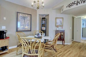 A dining area with a glass-top table surrounded by wicker chairs, a chandelier, and framed artwork on the walls. - Home Away From Home Unit J (Surfside Beach)